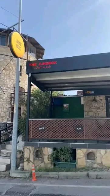 The Jukebox from the road by day: a stone building, the raised terrace with darts and pool signs, the red neon sign and a round yellow Jukebox sign on a pole
